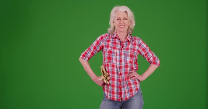 Portrait of proud senior woman standing with hands on hips holding gardening gloves on greenscreen. Friendly elder woman in flannel shirt smiling at camera on green screen to be keyed or composited. 