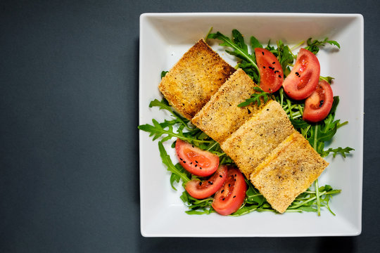 Salad of fried tofu with tomatoes and rocula. Homemade vegetarian salad in a white plate on a black background. A healthy Asian diet vegan vegetarian salad