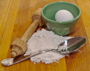 Utensils for baking, a spoon, a green bowl and a wooden rolling pin along with flour and an egg on a wooden cutting board 