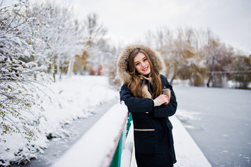 Fototapeta na wymiar Beautiful brunette girl in winter warm clothing. Model on winter jacket against frozen lake at park.