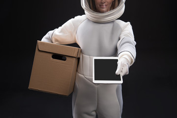 Sign it here. Pleasant spacewoman courier wearing helmet and protective suit is standing and keeping cardboard box while holding out digital tablet in hand. Selective focus. Space delivery concept