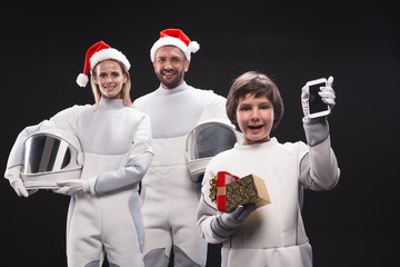 Portrait of happy boy spaceman is holding opened christmas box while rising hand with new phone and looking at camera with joy. His parent astronauts in red hats are posing in background. Isolated © Yakobchuk Olena