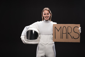 Migration to red planet. Portrait of delighted spacewoman wearing full armor is looking at camera with joy while holding white helmet and mars sign. Isolated background. Resettlement concept