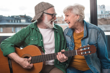 Happy time together. Mature man and woman sitting at window while looking at each other and playing guitar