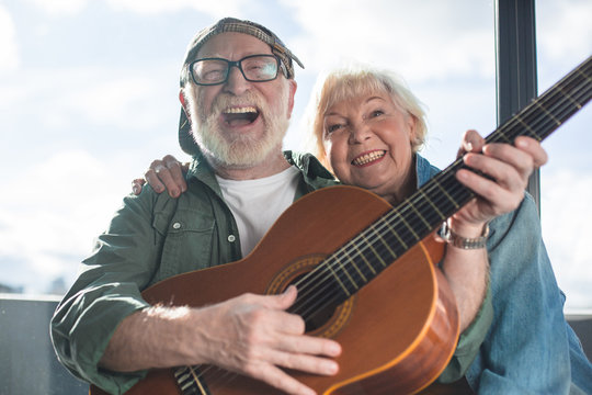 True happiness. Portrait of two sincere pensioners sitting together. Woman is cuddling man in cap while he is playing - Powered by Adobe