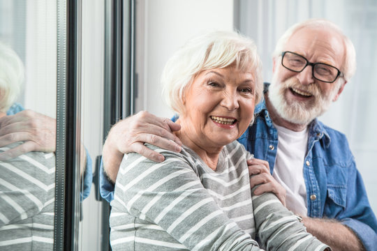 Amazing retirement. Waist up portrait of two old cheerful pensioners smiling at camera. Husband is tenderly hugging wife while sitting at window