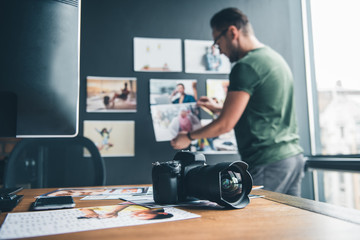 Focus on camera and photos locating on desk in office. Side view unshaven serene man putting pictures on background. Creativity and occupation concept