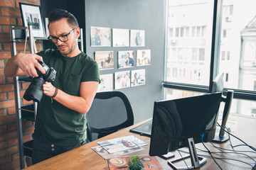 Portrait of serene bearded man watching at camera while holding it in hand. He stand near desk in office. Work concept