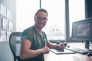 Portrait of happy unshaven photographer writing in electronic tablet while sitting at table in room. Occupation concept