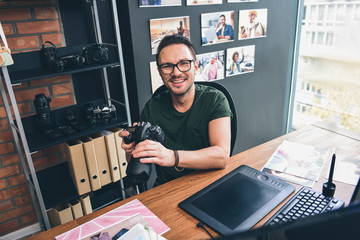 Portrait of beaming unshaven man keeping appliance for taking photos while sitting at table in room. Occupation concept