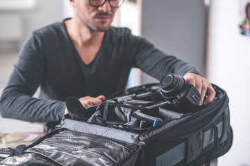 Close up male hand putting camera into big bag with other gadgets for images. Profession concept