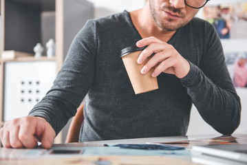 Close up hand of unshaven serious designer keeping mug of coffee. He looking at images in apartment. Work and hobby concept