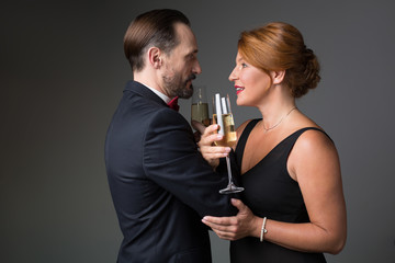 Side view profile of affectionate middle-aged married couple are drinking champagne while touching their arms to each other. They are looking into eyes with love and smiling. Isolated