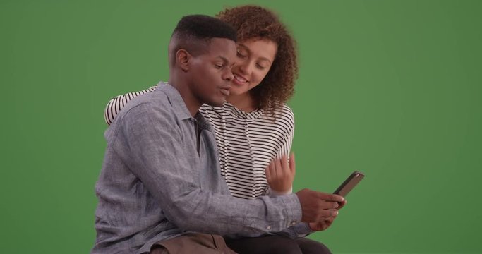 Young black couple sit on a bench looking at their smart phone on green screen. On green screen to be keyed or composited. 