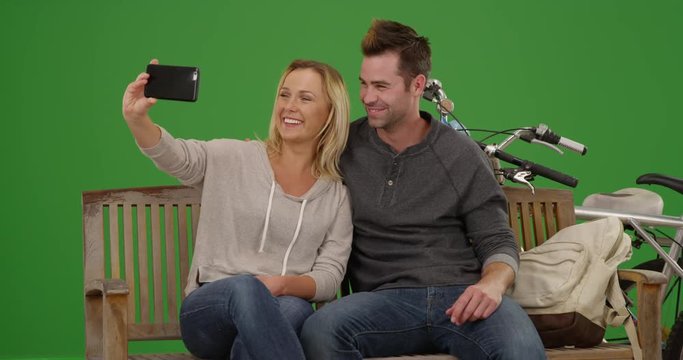 Hipster couple taking selfies on a bench with their bikes on green screen. On green screen to be keyed or composited. 