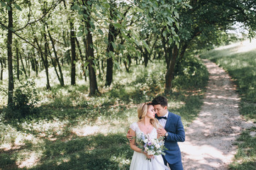 Fototapeta premium The groom tenderly embraces the bride by the shoulders. The bride is holding a bouquet. Wedding walk in the forest.