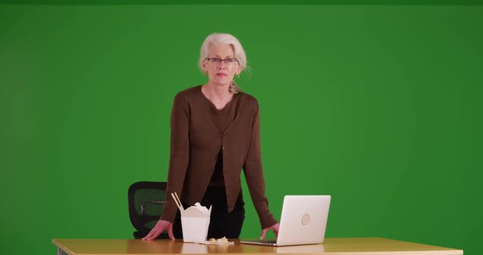 Portrait of an older woman looking at the camera with Chinese food and a laptop on her desk on greenscreen. Woman staring at the camera next to noodles and a computer. 