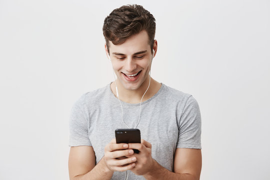 Handsome caucasian young male with dark hair and blue eyes holding mobile phone, smiling with teeth, typing message to his girlfriend, wearing earphones. People, lifestyle and modern communication.