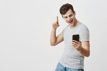 Handsome male can`t stop smiling as listens to jokes with earphones, pointing with forefinger...