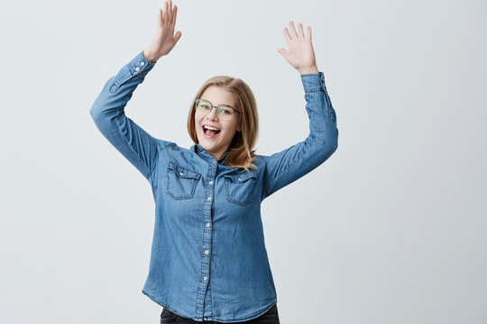 Overjoyed female wears danim shirt, stylish eyewear, gestures in excitement, exclaims loudly in astonishment, being glad to meet old friend, raises arms up. Blonde female rejoices success at work.
