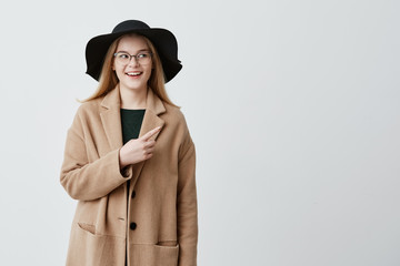 Fototapeta premium Beautiful smiling woman in coat over green sweater and eyeglasses pointing at blank white wall while demonstrating something. Cheerful young female indicating with fore finger on gray background.