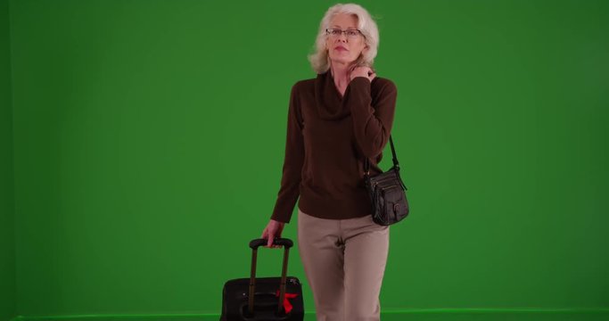 Elderly woman walking toward the camera with her suitcase and purse on greenscreen. Senior walking looking around with baggage on green screen to be keyed or composited.