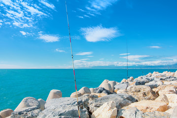 Fishing rods poles standing on big stones at the pier on sunny day, sea ocean lake blue transparent water under beautiful blue sky at the background.