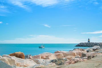 Panoramic seascape of Mediterranean sea with a lonely fishing boat and a small black and yellow lighthouse at Benalmadena port, Malaga province, Spain. Big stones at sandy bay beach at foreground.