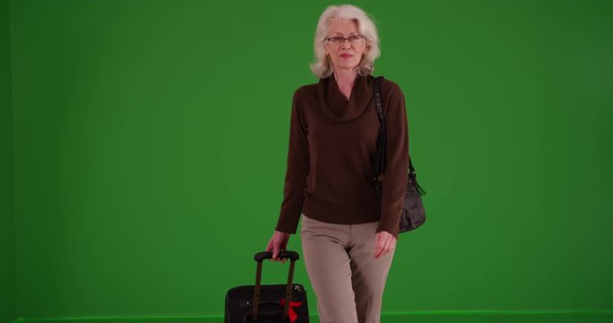 Elderly woman happily walking toward the camera with her suitcase and purse on greenscreen. Senior walking looking around with a smile with baggage on green screen to be keyed or composited.