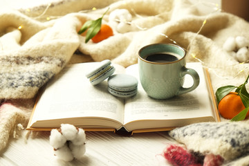 Close up blue coffee cup on the open book with cotton flower, clementine mandarin, french macaroons, plaid, and glowing christmas lights on the window sill. Cosiness, holiday morning comfort concept