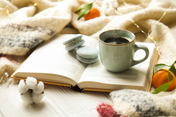 Obraz na płótnie Canvas Close up blue coffee cup on the open book with cotton flower, clementine mandarin, french macaroons, plaid, and glowing christmas lights on the window sill. Cosiness, holiday morning comfort concept
