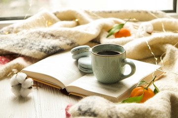 Close up blue coffee cup on the open book with cotton flower, clementine mandarin, french macaroons, plaid, and glowing christmas lights on the window sill. Cosiness, holiday morning comfort concept