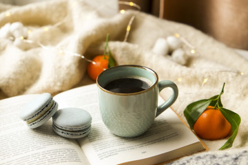 Close up blue coffee cup on the open book with cotton flower, clementine mandarin, french macaroons, plaid, and glowing christmas lights on the window sill. Cosiness, holiday morning comfort concept