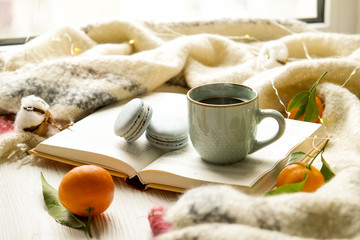 Close up blue coffee cup on the open book with cotton flower, clementine mandarin, french macaroons, plaid, and glowing christmas lights on the window sill. Cosiness, holiday morning comfort concept