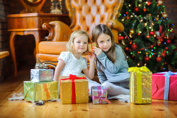 beautiful sisters sit on the floor near the Christmas tree and take gifts.