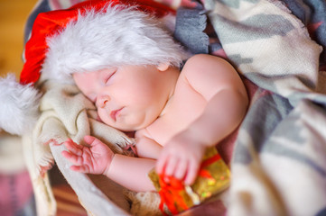 beautiful baby in Santa Claus hat lying asleep in basket, hugging a small box with a gift.