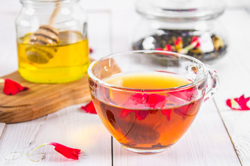 A cup of tea, in the background of a bank of honey and a jar with a black herbal floral tea on a white wooden table.