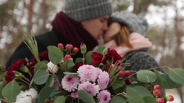 Newlyweds groom and bride hug kiss and warm each other in snowy pine forest during snowfall in slow motion. . Young couple in winter wood. Woman with bouquet. Valentine's Day and Christmas concept.
