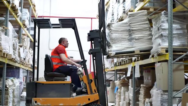 Warehouse worker driver in uniform moving cardboard boxes by forklift stacker loader. Forklift pallet with boxes in large modern warehouse