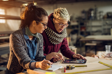 Close up view of hardworking focused professional motivated engineer woman working with a customer while choosing a product on a tablet in the sunny fabric workshop.