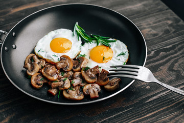 Vegetarian breakfast with fried eggs and mushrooms decorated with a rosemary sprig in a frying pan on a wooden table