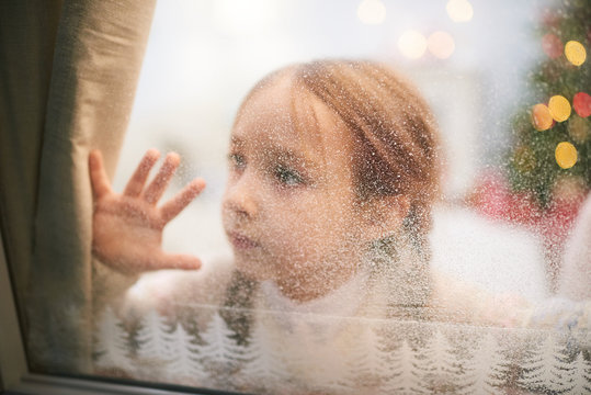 Pretty little girl wearing knitted sweater looking out window covered with frost while waiting for Santa Claus, portrait shot - Powered by Adobe
