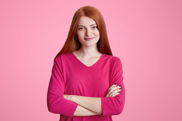 Obraz premium Cute freckled female with long ginger hair stands crossed hands, wears pink sweater, being pleased to meet with friends, isolated over pink background. Pleased red haired woman poses indoor.