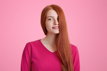 Fototapeta premium Portrait of pleased smiling young female with long ginger hair covering her face, models for well known magazine, isolated over pink background. Joyful woman with charming smile. Beauty concept