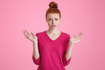 Unhappy displeased female model with hair bun, shrugs shoulders in bewilderment, makes questioning gesture, being uncertain, has confused clueless look. Gloomy ginger young woman poses indoor