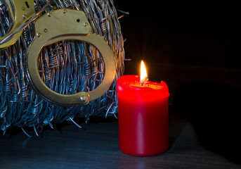 eine rote Kerze leuchtet vor Stacheldraht und Handschellen