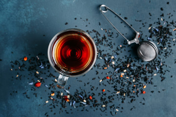 Glass cup of black tea, tea stainer and sprinkled tea leaves on dark background