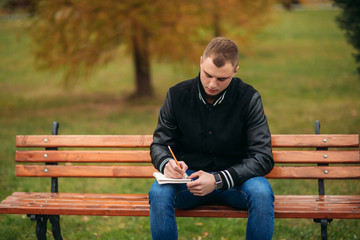Fototapeta premium A student in a black jacket sits in a park on a bench writes down his thoughts in a notebook. Handsome boy