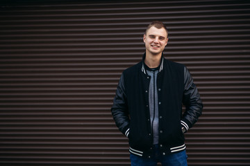 A young guy in a black jacket against a background of dark striped walls posing and smiling at the photographer
