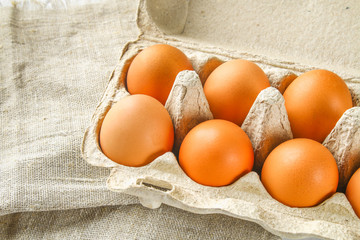 Raw brown chicken eggs in a cardboard tray with cells on sacking on a white wooden table. Ingredients for cooking.